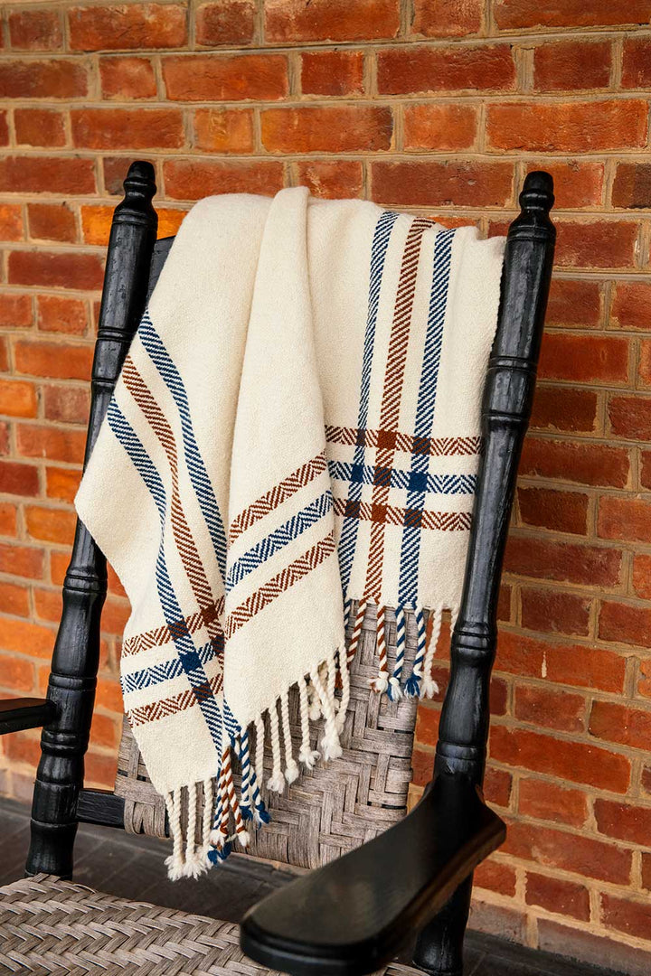 The Limited Edition Merino Wool Blanket, cream-colored with blue and brown plaid stripes and fringe, hangs over a black wooden rocking chair in front of a red brick wall.