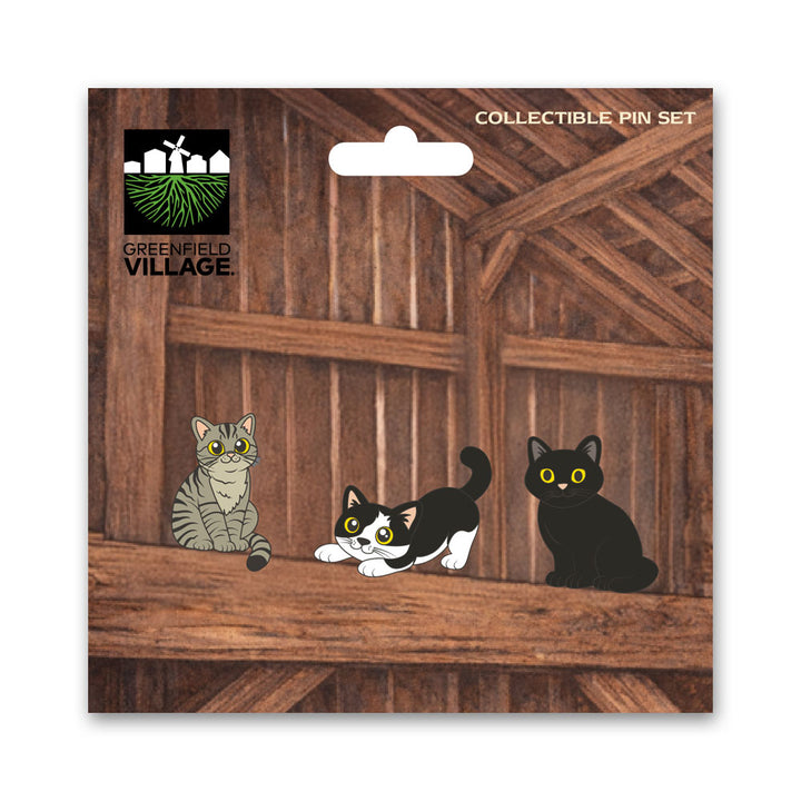 The Greenfield Village Barn Cats Pin Set features three cartoon cat pins—a gray tabby, a black and white, and a solid black cat—displayed on a wooden loft background with the Greenfield Village logo and the product name at the top.