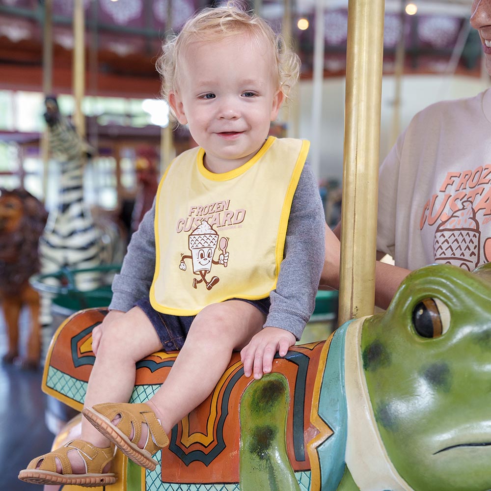 A smiling toddler in a Frozen Custard Bib sits on a green carousel frog at Greenfield Village, with brown sandals. An adult stands nearby, while a zebra carousel animal and frozen custard keychain are seen in the background.