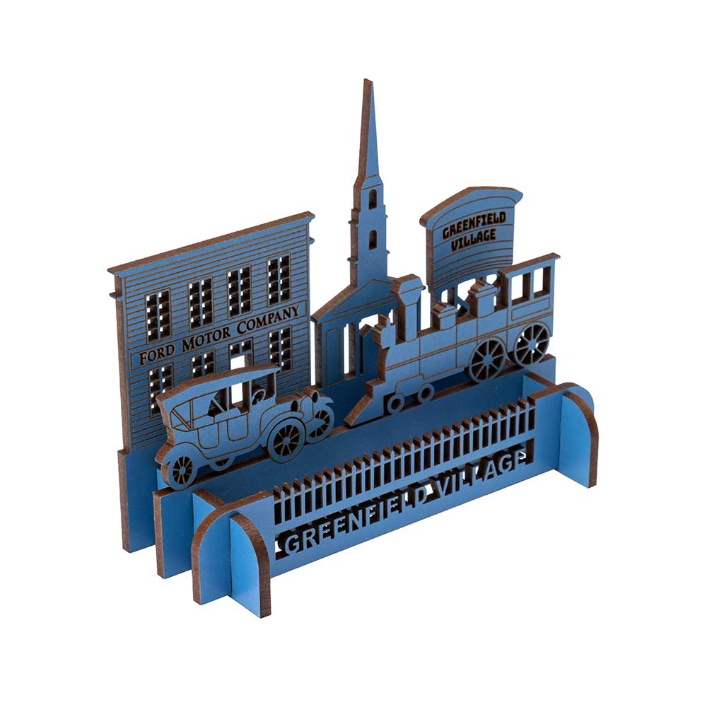 The Henry Ford® Greenfield Village 3D Wooden Puzzle features a Ford Model T, steam train, church steeple, and a Ford Motor Company building. Painted blue and brown, the base is labeled GREENFIELD VILLAGE.