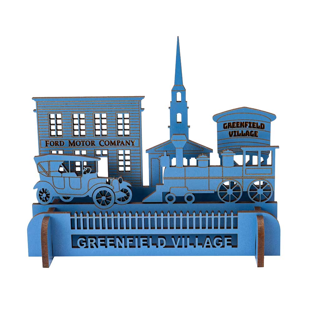 The Henry Ford® Greenfield Village 3D Wooden Puzzle is a blue, laser-cut set featuring a vintage car, steam locomotive, church with steeple, and buildings labeled Ford Motor Company and Greenfield Village.