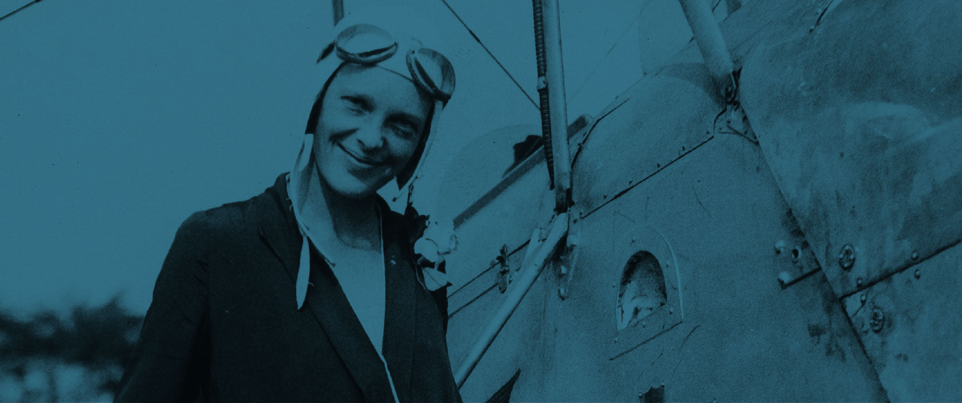 A person wearing aviator goggles and a flight cap smiles while standing next to a vintage airplane. The image has a blue tint.