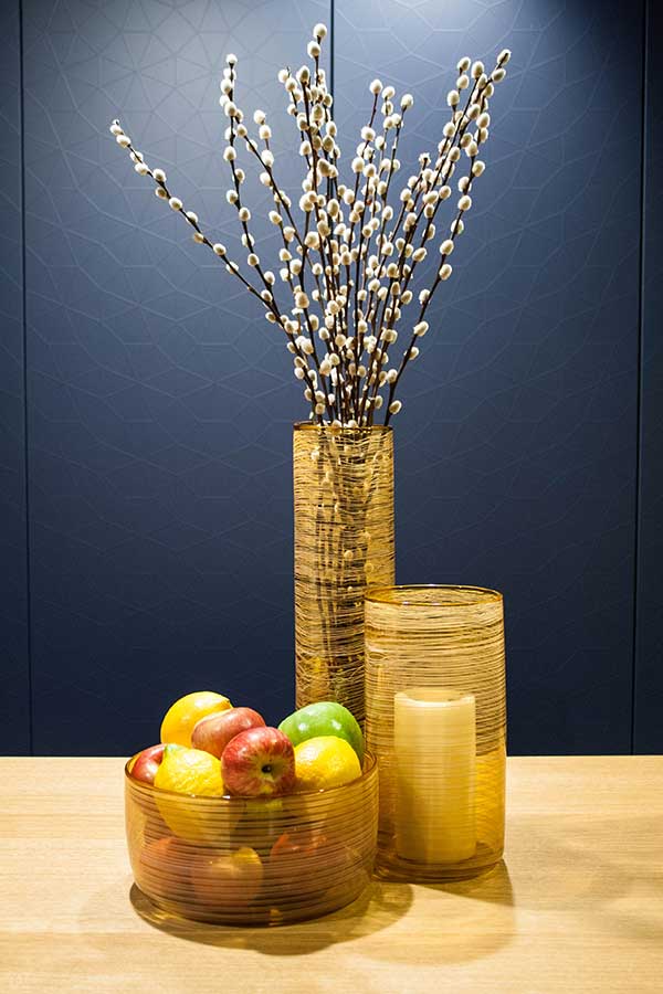 A still life features a modern Matassa Tall Cylinder Vase with pussy willow branches, a glass fruit bowl, and a cylindrical candle holder on wood, set against a dark blue background.