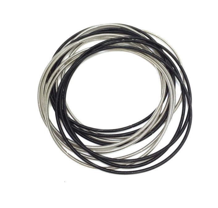 The Unbound Black and Silver Piano Wire Bracelet, featuring coiled strands in black and silver, is displayed in an overlapping circular pattern on a white background.
