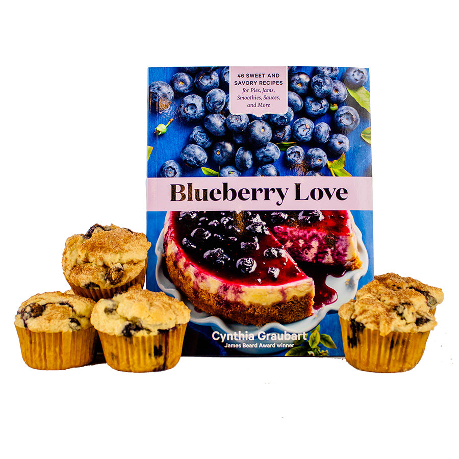Four blueberry muffins sit in front of the cookbook "Blueberry Love," which features blueberry recipes and a cover with fresh blueberries and a blueberry dessert.