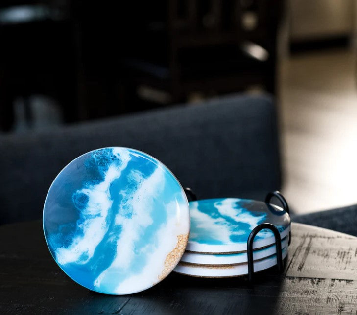 Ceramic & Resin Coasters - Ocean Vibes feature blue, white, and tan wave designs, displayed in a black holder on a dark table—making them a unique gift for any occasion.