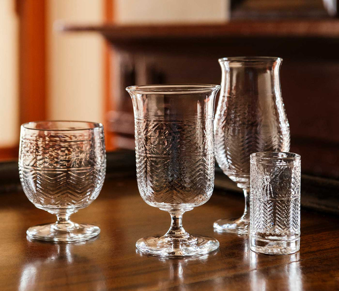 Four clear American Heritage Molded Tulip Glasses are arranged on a polished wooden table, highlighting their intricate patterns against a softly blurred background.