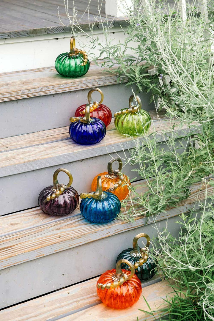 The Small Glass Pumpkin - Aqua is a hand blown collectible keepsake inspired by Greenfield Village, featuring vibrant aqua color and unique artistry—perfect for display beside plants or on wooden outdoor steps.