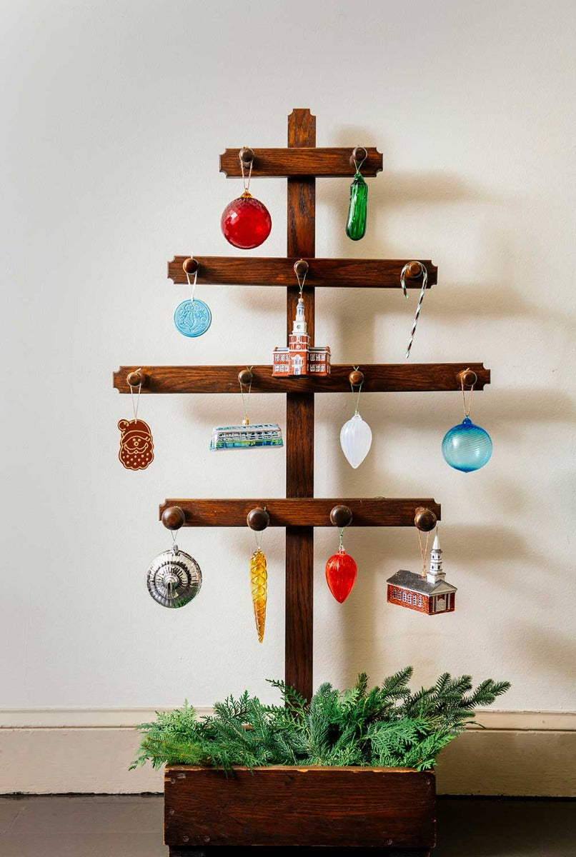 A minimalist wooden Christmas tree with horizontal branches displays a Hand Blown Glass Pickle Ornament and stands in a box with green pine at the base, set against a plain white wall.