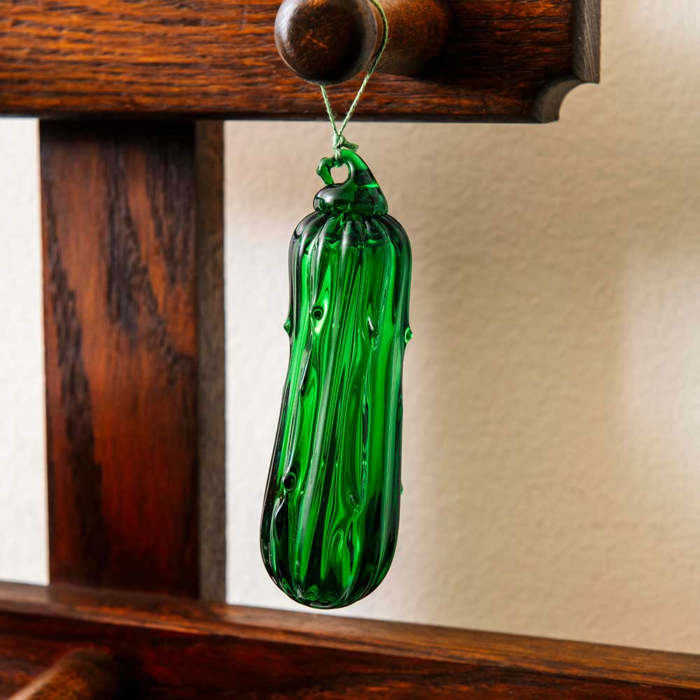 A Hand Blown Glass Pickle Ornament, cherished in family traditions for bringing good luck, hangs by a string from a wooden rack against a light-colored wall.