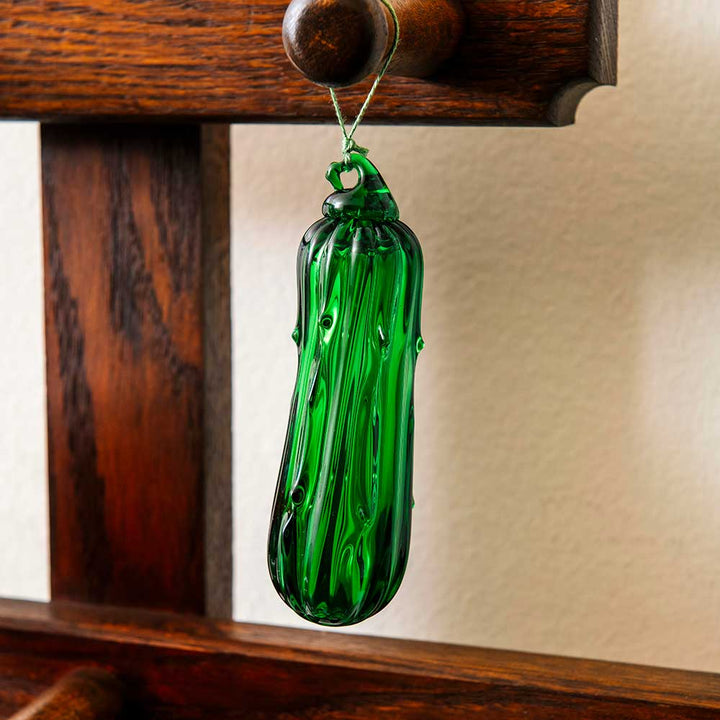 A Hand Blown Glass Pickle Ornament, cherished in family traditions for bringing good luck, hangs by a string from a wooden rack against a light-colored wall.
