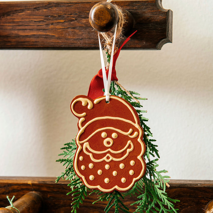 Handcrafted, the Redware Santa Ornament features a Santa-shaped design with white icing details and hangs from a ribbon and greenery on a wooden hook, reminiscent of ornaments seen in Greenfield Village.