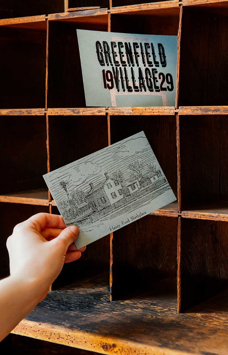 A hand places the Henry Ford Birthplace Postcard, featuring antique wood type, into a mail cubby labeled GREENFIELD VILLAGE 29.