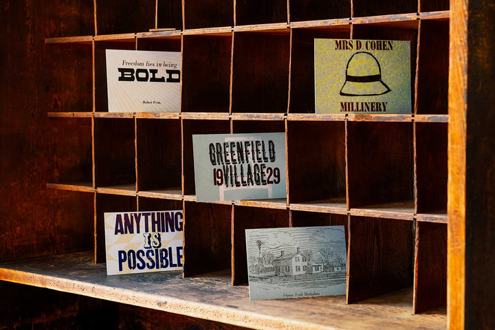 A wooden cubby shelf displays vintage-style cards, including the “Freedom Lies in Being Bold” postcard, “MRS D COHEN MILLINERY,” “GREENFIELD VILLAGE 29,” and a Robert Frost postcard with a building sketch.