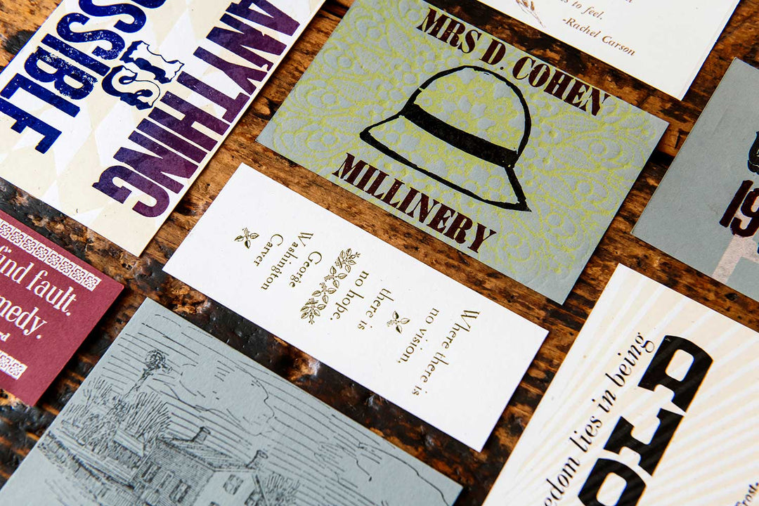 A close-up of vintage-style cards on a marbled surface, including the Mrs. D Cohen Millinery Postcard with a pith helmet, and others featuring bold fonts made from antique wood and metal type.