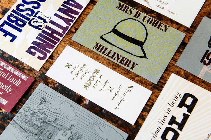A close-up of vintage-style cards on a marbled surface, including the Mrs. D Cohen Millinery Postcard with a pith helmet, and others featuring bold fonts made from antique wood and metal type.