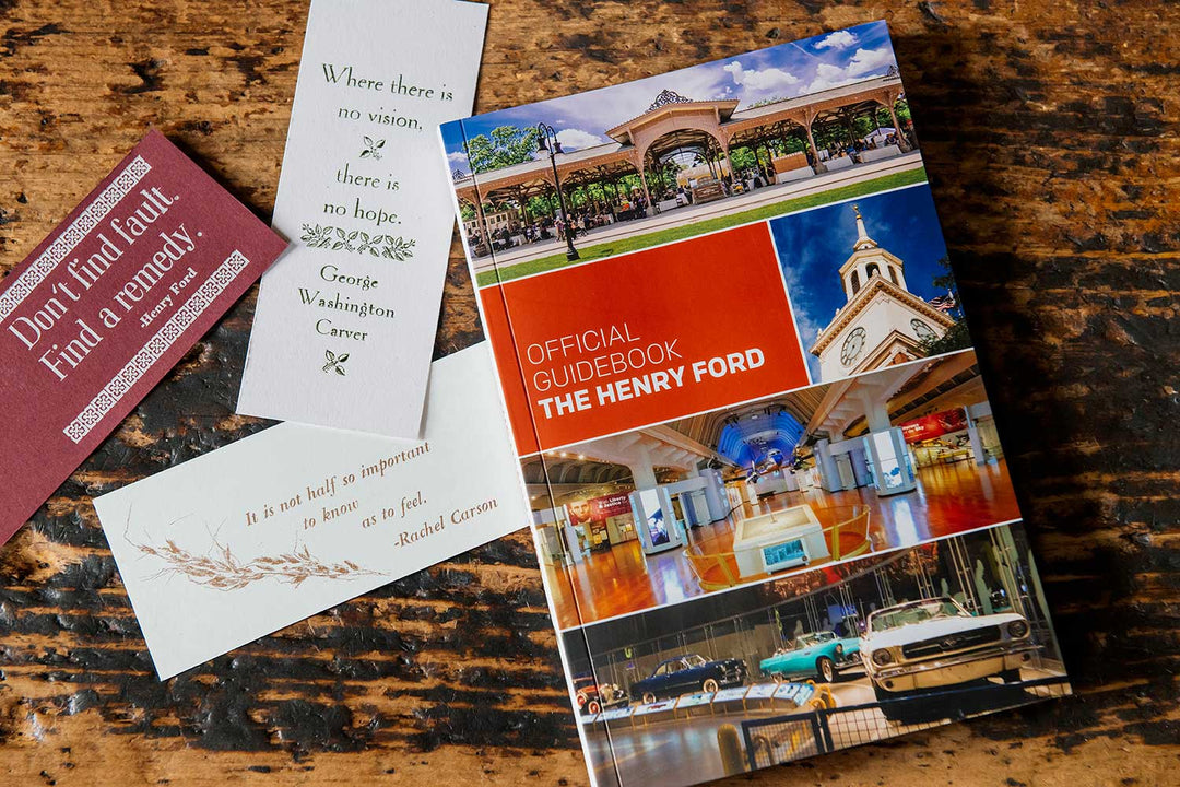 The Henry Ford Official Guidebook rests on a wooden surface, accompanied by three inspirational bookmarks. The cover displays exhibits from the Henry Ford Museum and historic Greenfield Village buildings.