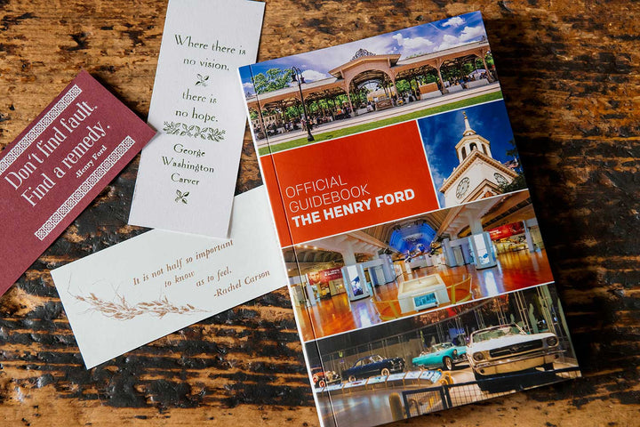 The Henry Ford Official Guidebook rests on a wooden surface, accompanied by three inspirational bookmarks. The cover displays exhibits from the Henry Ford Museum and historic Greenfield Village buildings.