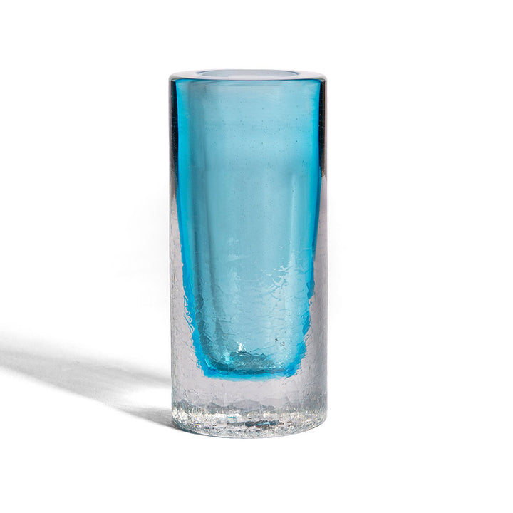 The Crackle Vase Tall - Blue features a cylindrical shape with a textured clear exterior and smooth translucent blue interior, echoing hand-blown glass artistry. It stands upright on a white surface.