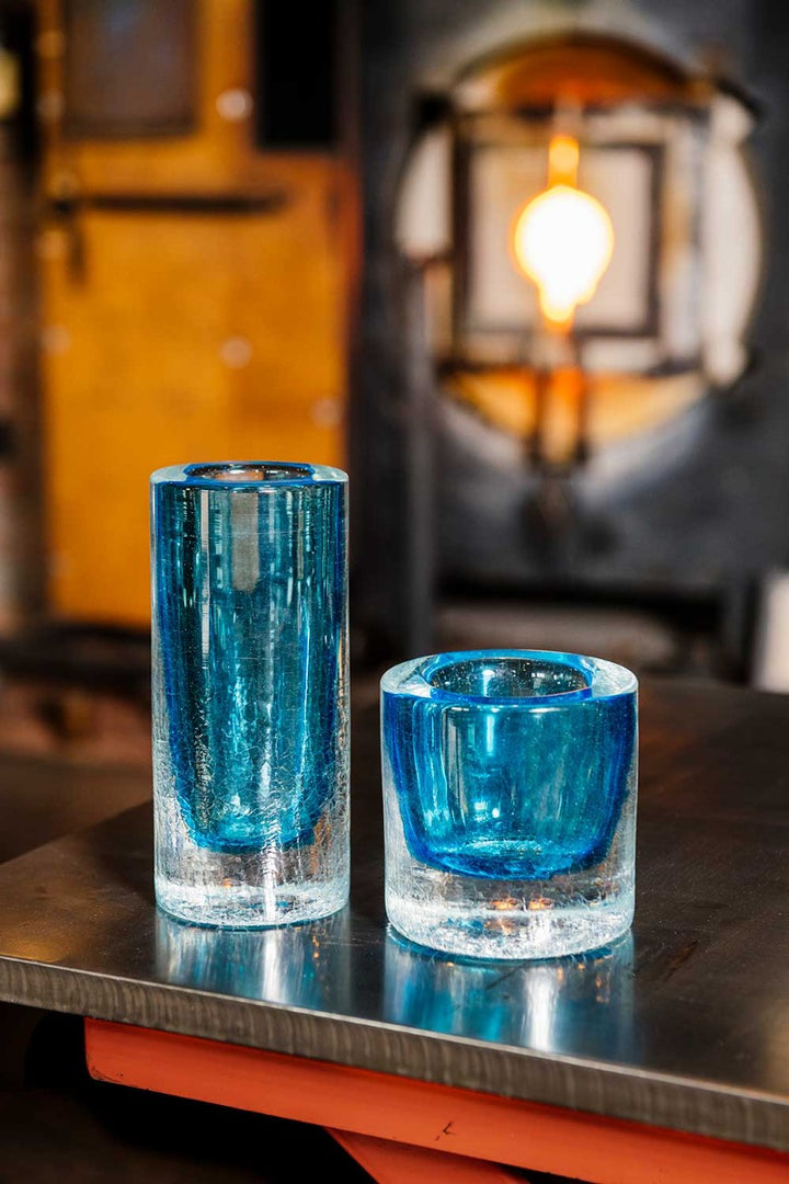 Two Crackle Vase Tall - Blue pieces with thick, clear bases sit on a metal surface. Behind them, the glowing glass furnace highlights the artistry of hand-blown glass in this vibrant workshop.