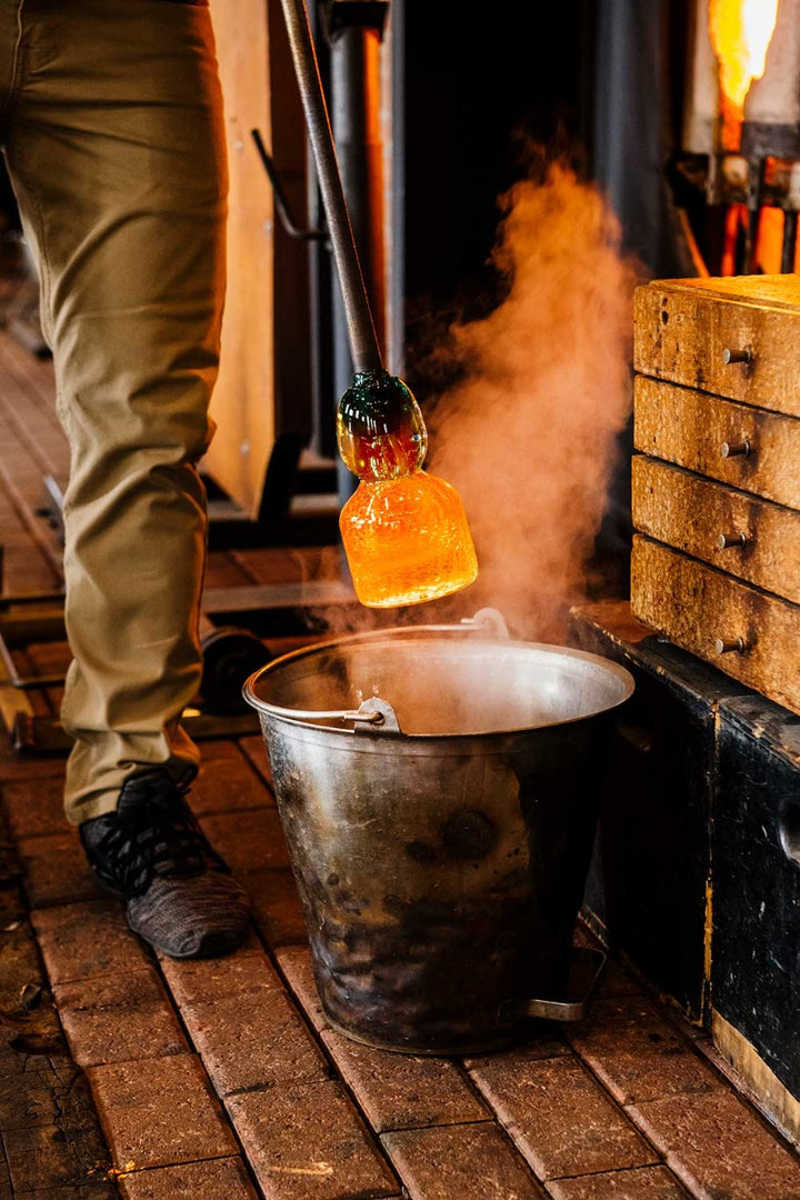 A person lowers the Crackle Vase Tall - Blue, a glowing hand-blown glass piece, into a metal bucket in a workshop with brick flooring and industrial equipment, creating a burst of steam.
