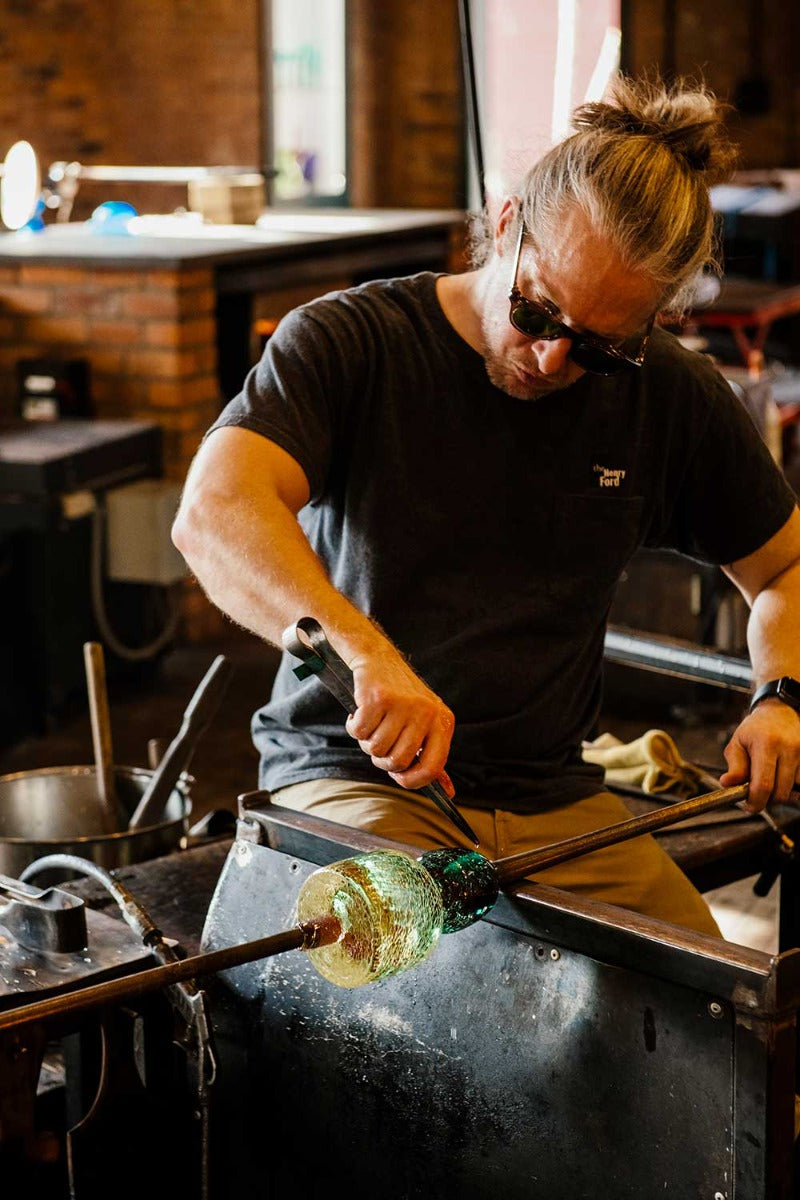 Wearing a bun and sunglasses, an artisan shapes the Crackle Vase Tall - Blue at a workshop table, using metal tools with skilled focus, capturing the artistry of traditional Murano glassmaking.