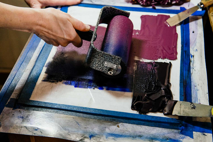 A hand rolls purple and black ink onto a taped surface with metal spatulas nearby, prepping materials to hand print the "Freedom Lies in Being Bold Postcard" using antique wood and metal type.