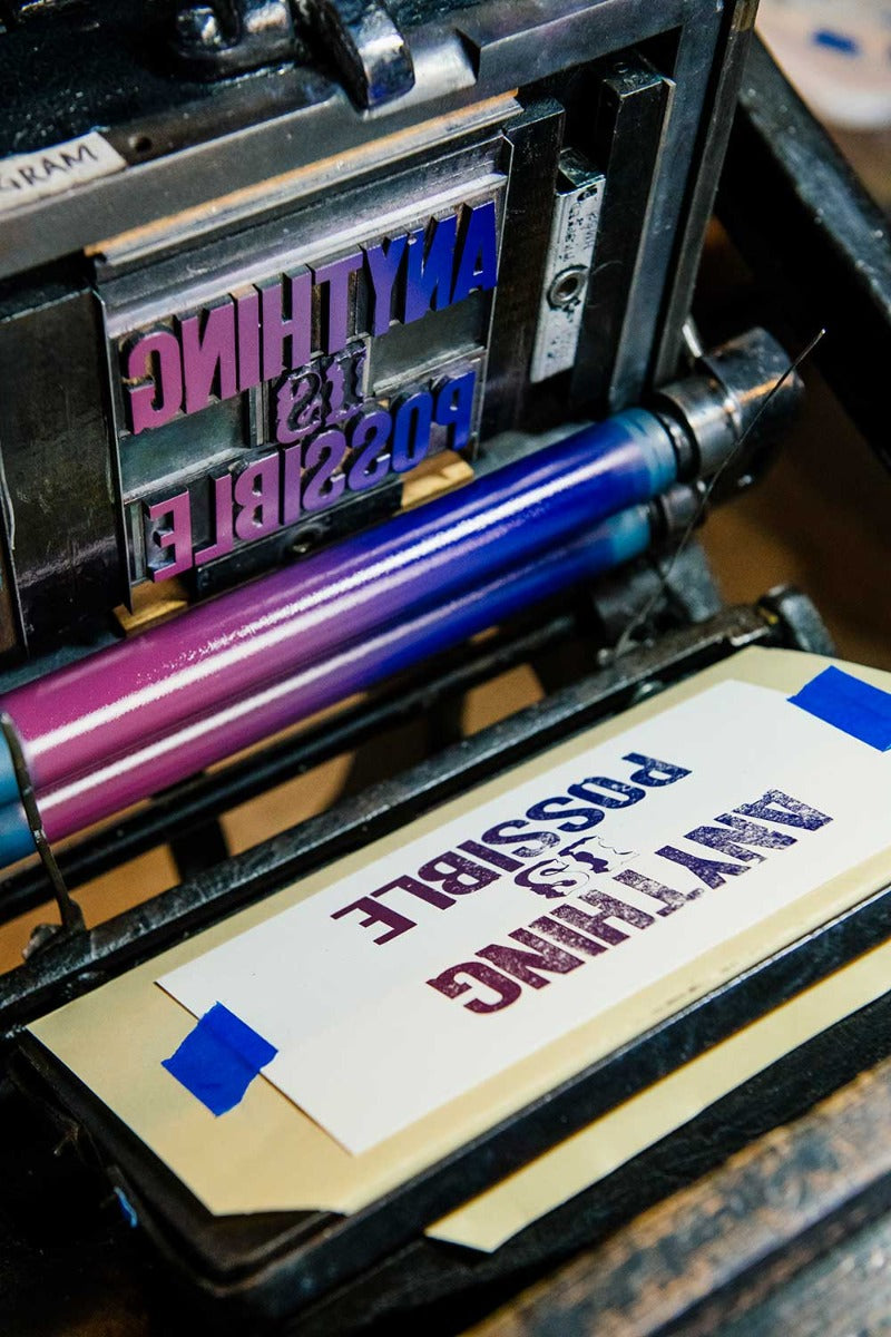 A close-up of the "Anything is Possible Postcard" being printed in purple and black ink with antique wood type, artisan techniques, and reversed text visible on the letterpress plate above.