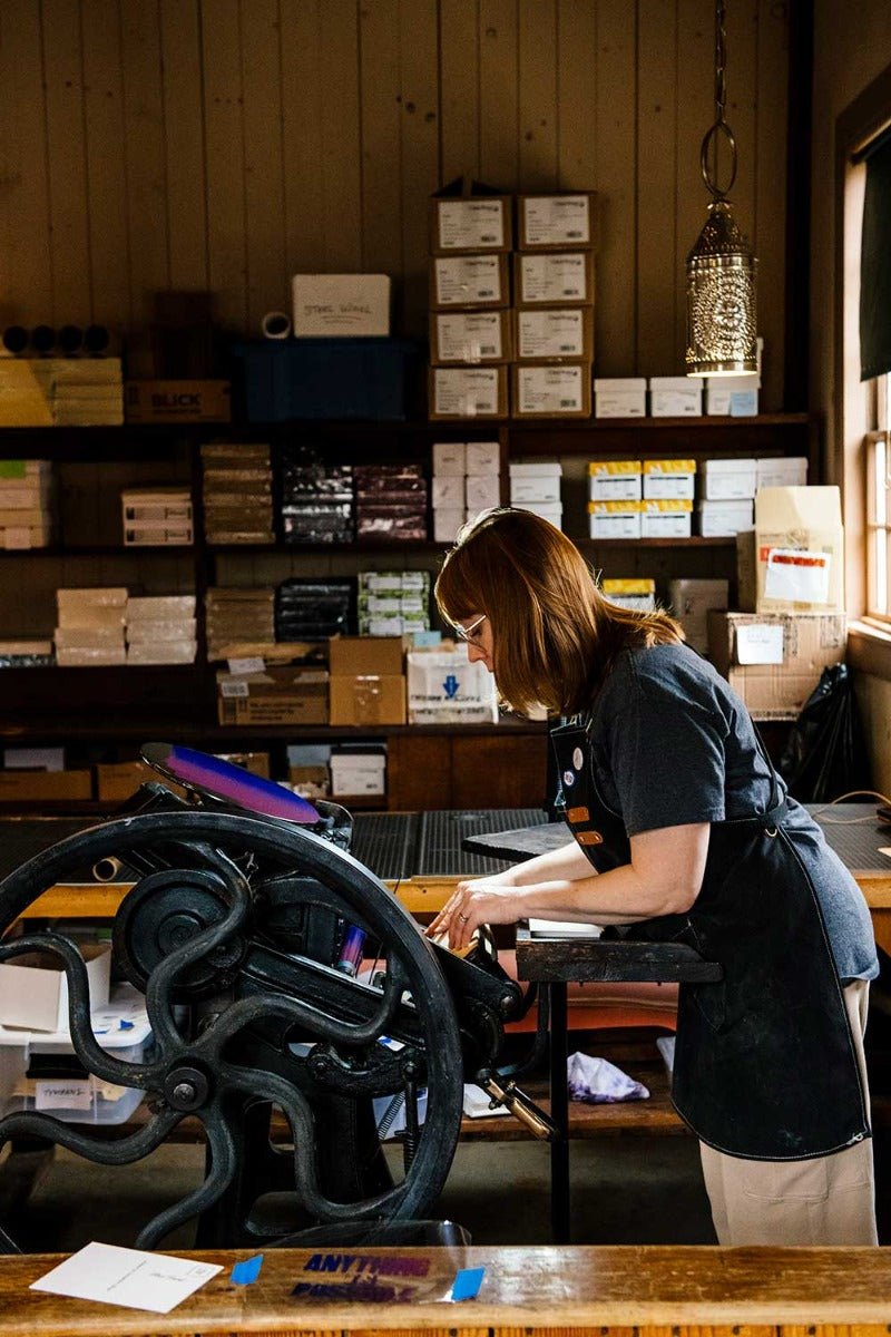 A woman in glasses and an apron operates a vintage printing press, crafting the Henry Ford Birthplace Postcard with antique wood type in a cozy workshop lined with shelves of boxes and paper supplies.
