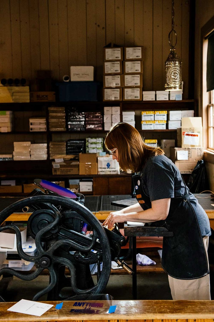 A woman in glasses and an apron operates a vintage printing press, crafting the Henry Ford Birthplace Postcard with antique wood type in a cozy workshop lined with shelves of boxes and paper supplies.