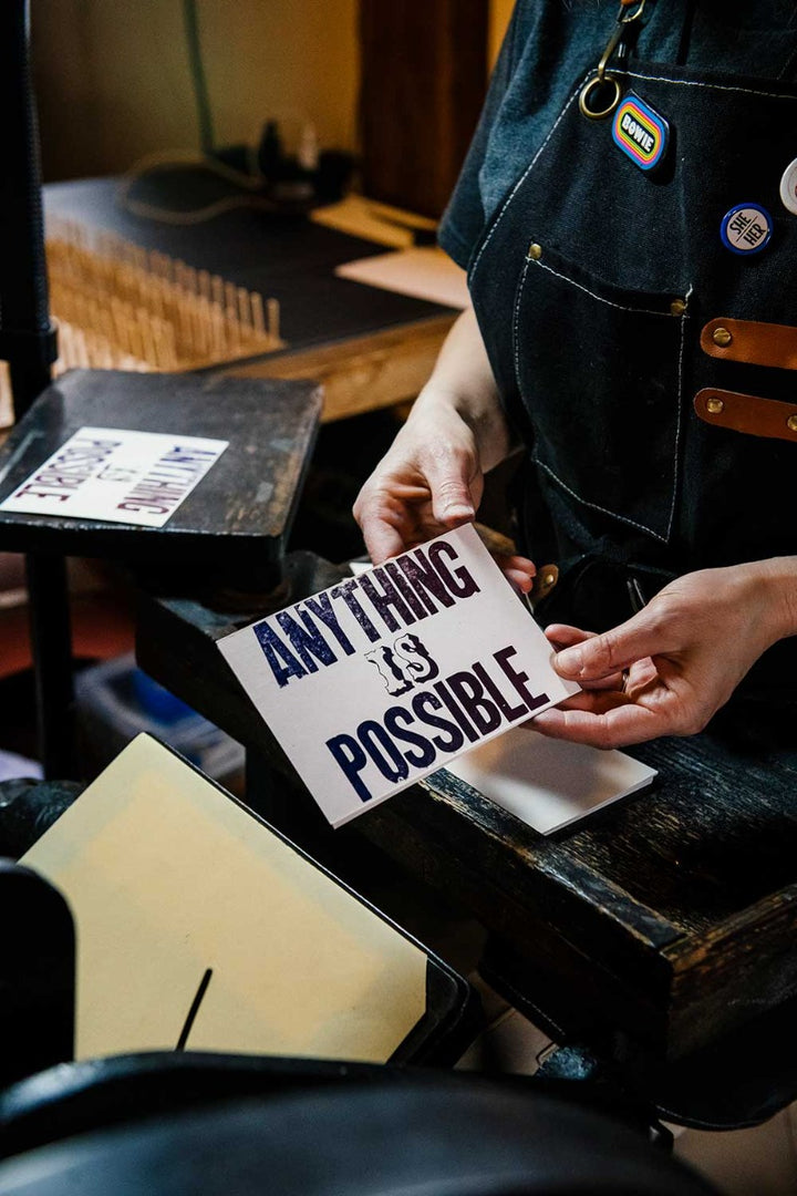 A person in a dark apron holds the "Anything is Possible Postcard," featuring bold hand-printed letterpress text, surrounded by antique wood type and artisan printing techniques in a workshop setting.
