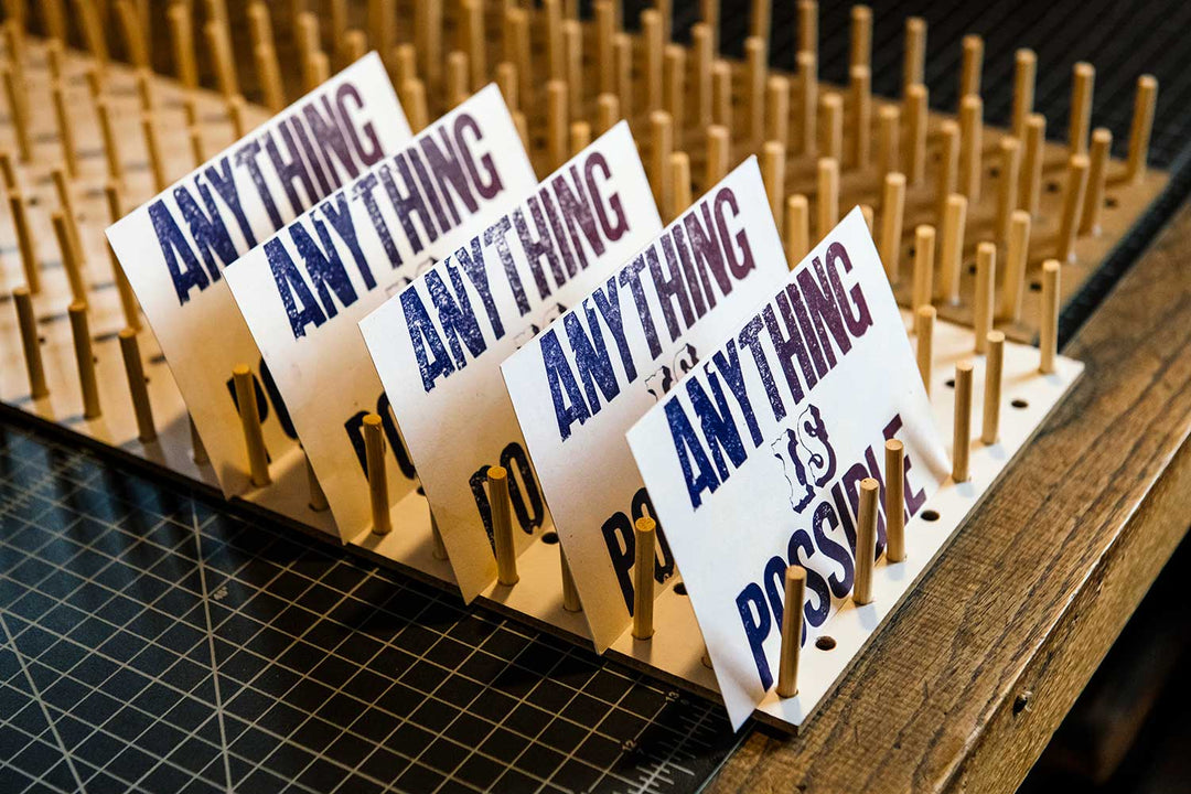 Four Anything is Possible Postcards are displayed upright on a wooden pegboard, highlighting artisan letterpress printing techniques above a workspace with a cutting mat and grid pattern.