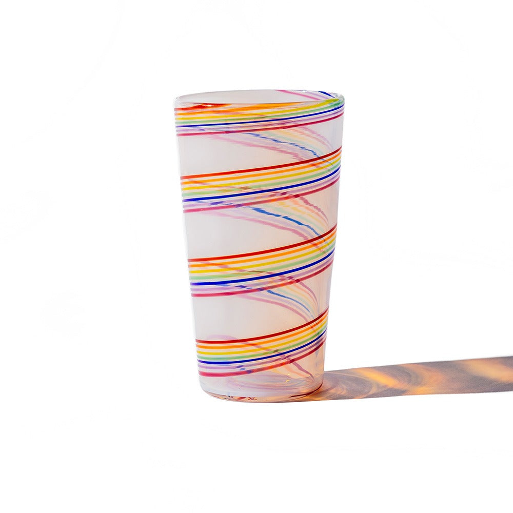 The Rainbow Cane Cup, a tall opaline glass with a white base and swirling lines in orange, blue, yellow, red, and purple, casts a multicolored shadow on a white background.