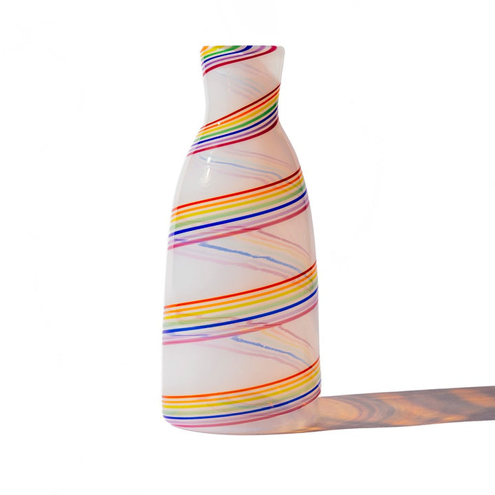 The Rainbow Cane Carafe is a white opaline glass bottle with a glossy finish and swirling rainbow-colored stripes, casting a soft shadow on a white background—perfect as a stunning centerpiece.