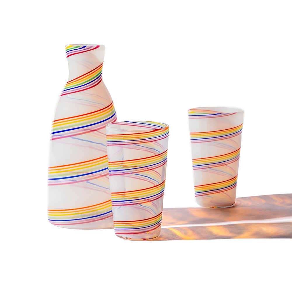 A Rainbow Cane Cup sake set—white bottle and two cups adorned with swirling, rainbow-hued lines—rests on a white surface, their shadows reflecting the same vibrant patterns.