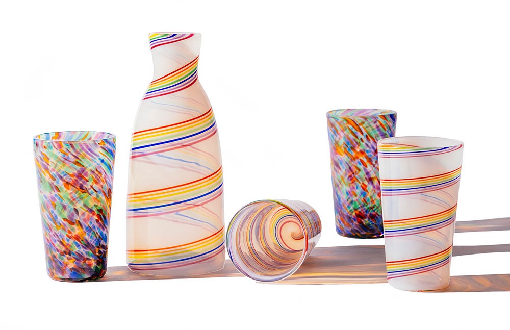 The Rainbow Cane Carafe and four colorful glasses with swirled and speckled designs are displayed on a white surface, with bright lighting casting lively shadows.