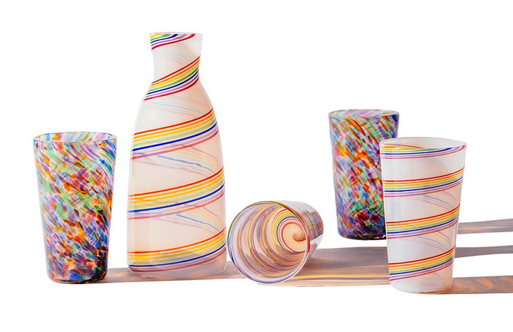 The Rainbow Cane Carafe and four colorful glasses with swirled and speckled designs are displayed on a white surface, with bright lighting casting lively shadows.