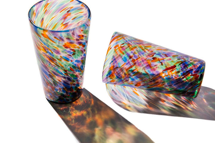Two Rainbow Frit Cups are displayed against a white background—one upright, one on its side—showcasing vibrant, rainbow-hued shadows that highlight the unique charm of each handblown Rainbow Frit Cup.