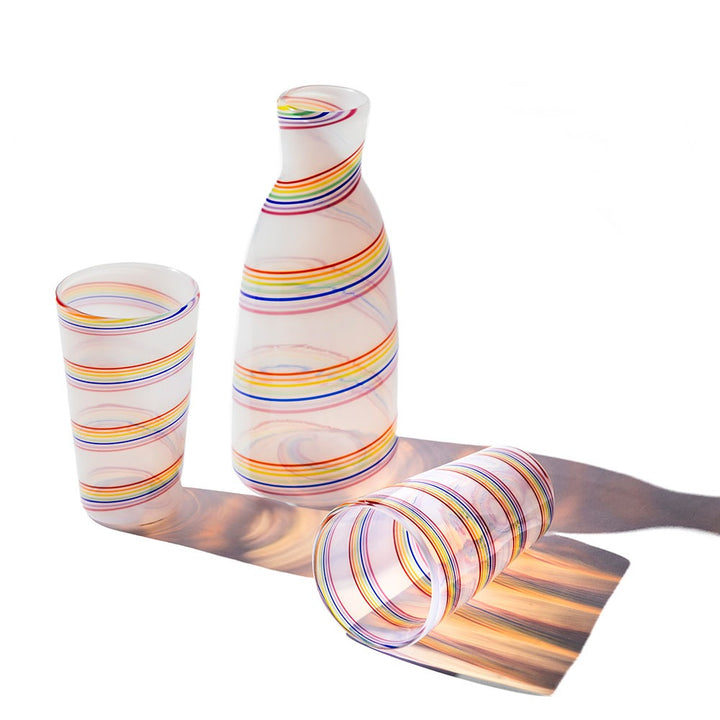 The Rainbow Cane Carafe with two opaline glass tumblers displays colorful swirling stripes; one tumbler stands upright and the other lies on its side, casting rainbow-hued shadows on a white surface.
