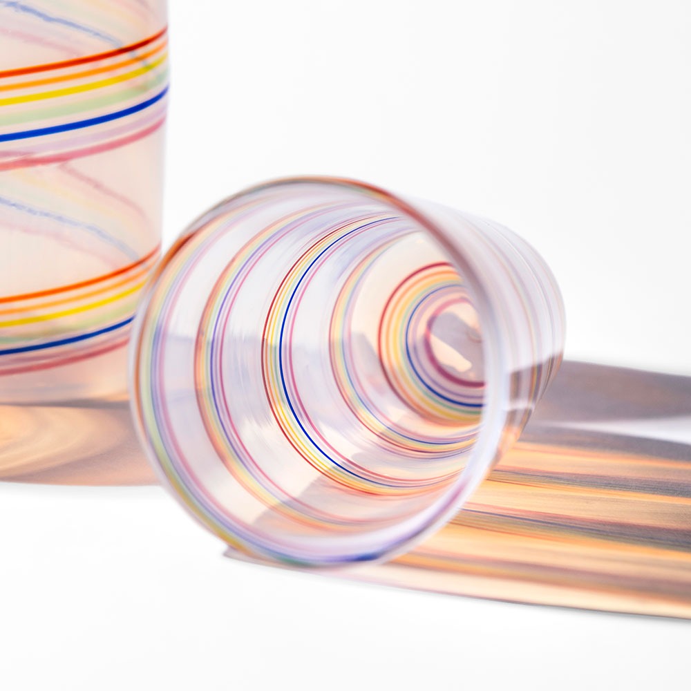 A Rainbow Cane Cup with vibrant swirling stripes rests on its side, casting a multicolored shadow on a white surface, while another Rainbow Cane Cup appears partially in the background.