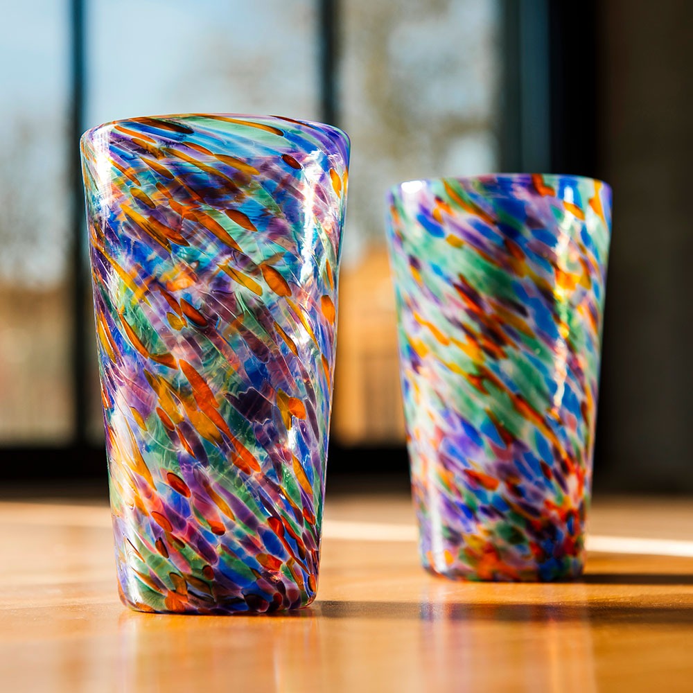 Two Rainbow Frit Cups, hand-blown with swirling blue, orange, green, purple, and white patterns, rest on a sunlit wooden surface against a softly blurred background.
