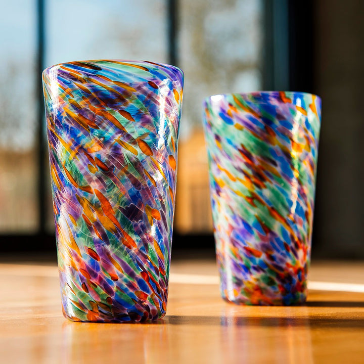 Two Rainbow Frit Cups, hand-blown with swirling blue, orange, green, purple, and white patterns, rest on a sunlit wooden surface against a softly blurred background.
