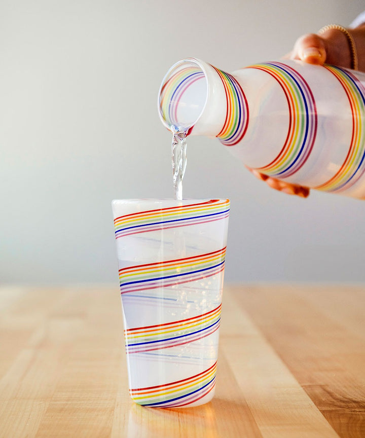 A hand pours water from a Rainbow Cane Cup pitcher into a matching glass, both made of vibrant rainbow glass, on a light wooden table with a softly blurred background.