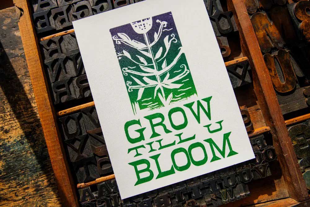 The Grow Till U Bloom Poster features a colorful linocut floral design with a stylized plant and bold green "GROW TILL U BLOOM" text, printed on soft cotton fiber paper. Displayed on a tray with wooden letter blocks.