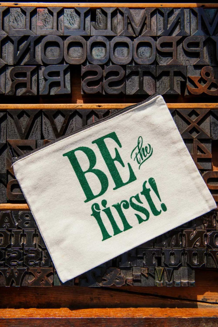 The Be the First Pouch is a cream-colored canvas zip-top pouch featuring green "BE the first!" text, displayed on vintage wooden letterpress blocks and inspired by the charm of the Greenfield Village Print Shop.