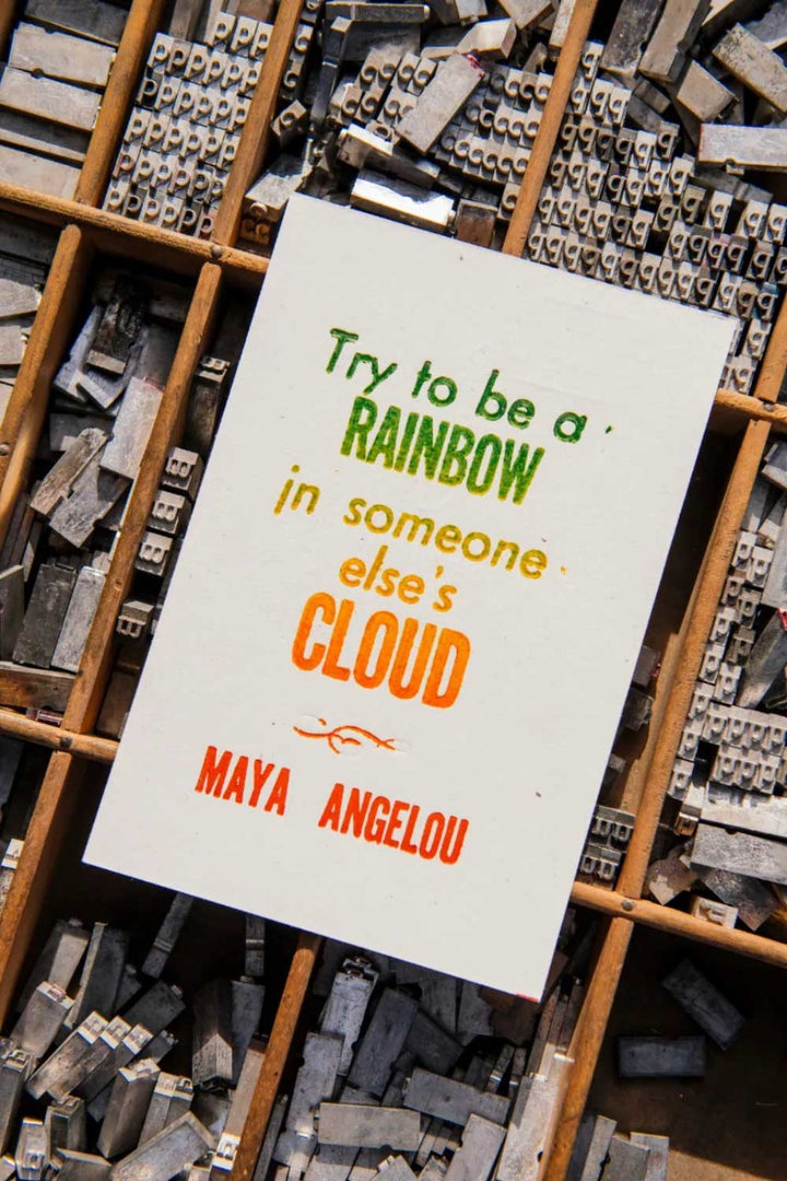 The Angelou Quote Postcard features vibrant rainbow text: "Try to be a rainbow in someone else's cloud," printed with rainbow roll technique and displayed on a wooden letterpress tray filled with metal type.