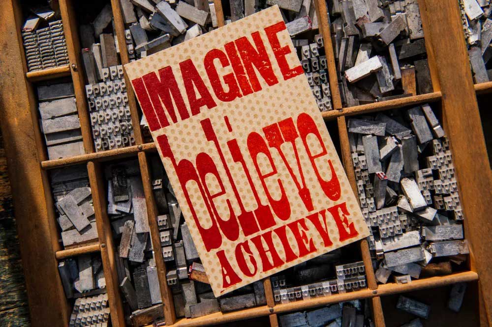 The Imagine Believe Achieve Postcard features bold red text and is displayed on a tray of vintage letterpress and antique wood type blocks.