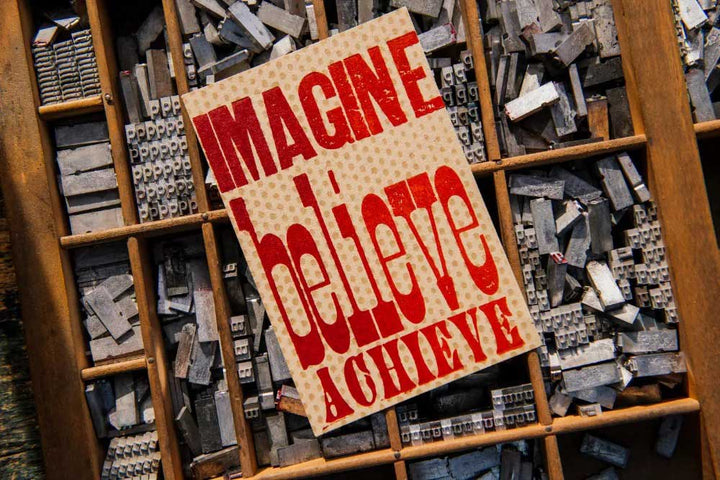 The Imagine Believe Achieve Postcard features bold red text and is displayed on a tray of vintage letterpress and antique wood type blocks.