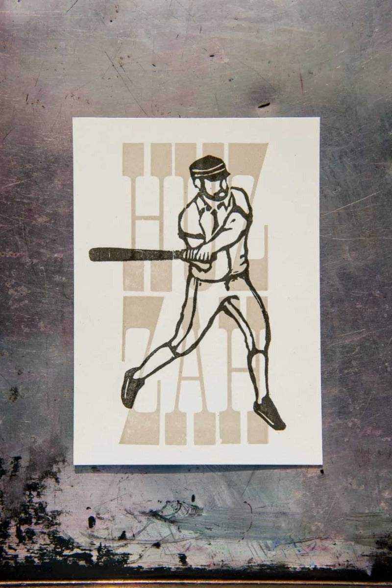 The Huzzah Mini Poster features a baseball player swinging a bat, with “HIT ZAG” in bold vintage type behind him. The artwork is centered on paper that sits atop a worn metallic surface, creating a nostalgic base ball vibe.
