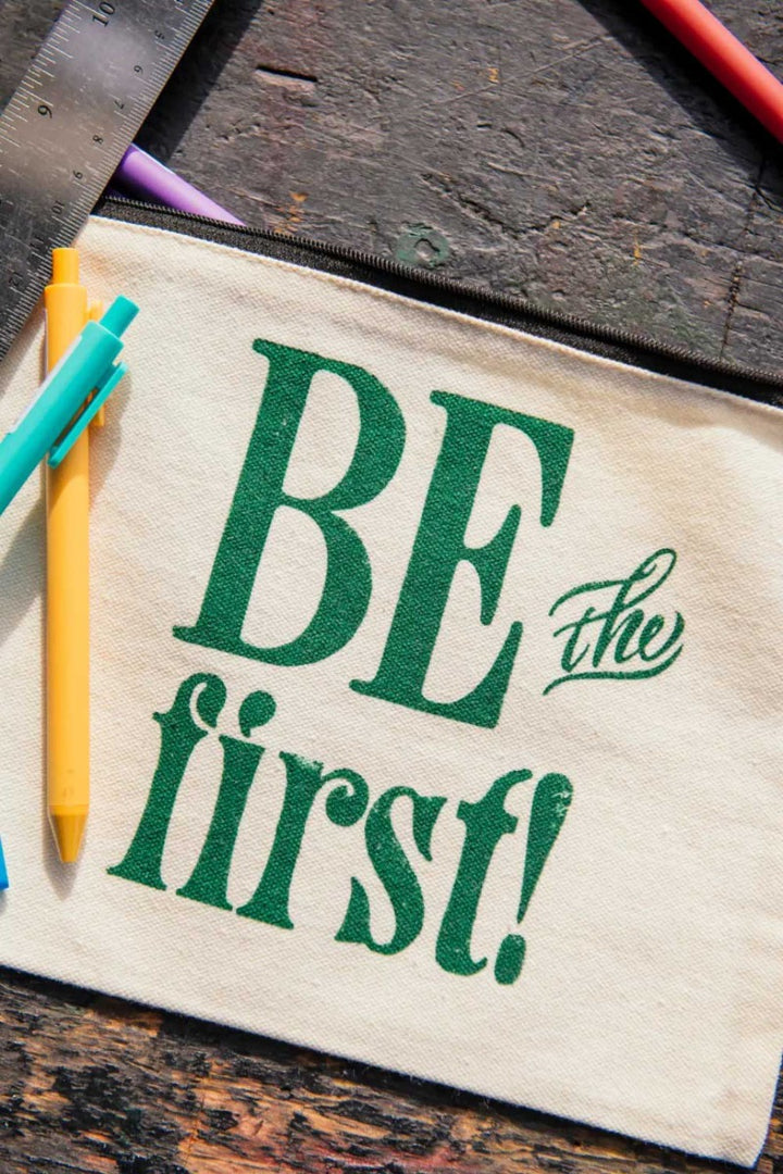 The Be the First Pouch, a cream-colored canvas zip-top bag featuring "BE the first!" in green letters, sits on a wooden surface with colorful pens and a metal ruler—perfect as a cotton art supply bag inspired by Greenfield Village Print Shop.